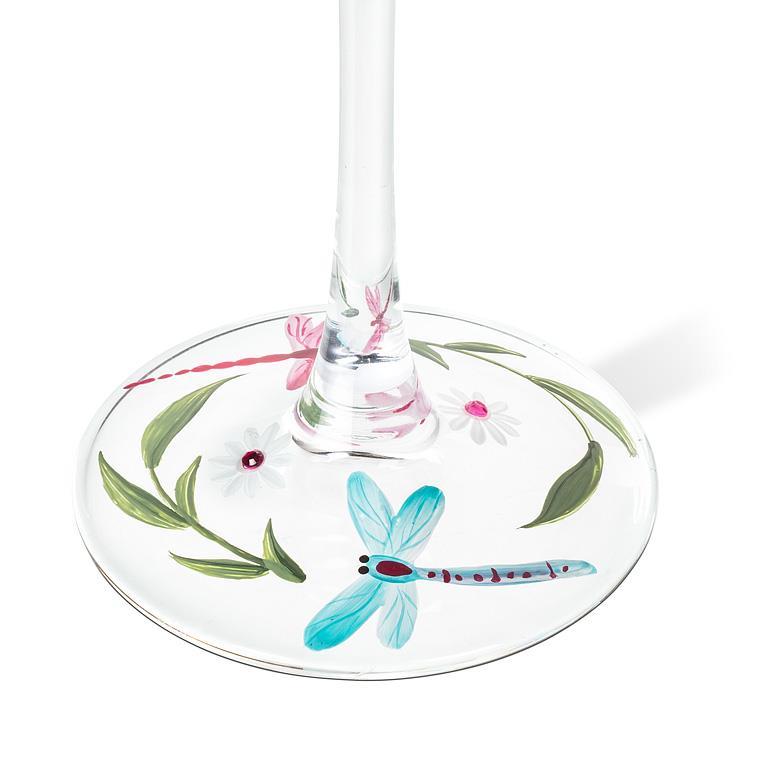Gros plan sur le pied d'un verre à vin transparent, illustré d'une libellule bleu turquoise, de feuilles vertes et de fleurs à centre rose.