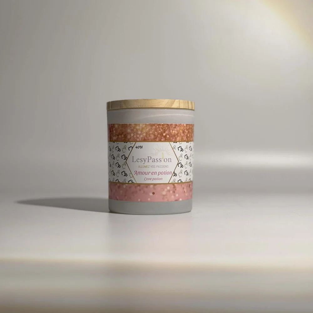 Bougie parfumée format 9 oz (40 h) en pot cylindrique gris très clair avec couvercle en bois, dotée d'une étiquette « LesyPassion - Amour en potion / Love potion » ornée de bandes pailletées rose poudré et or rose, sur fond studio neutre.