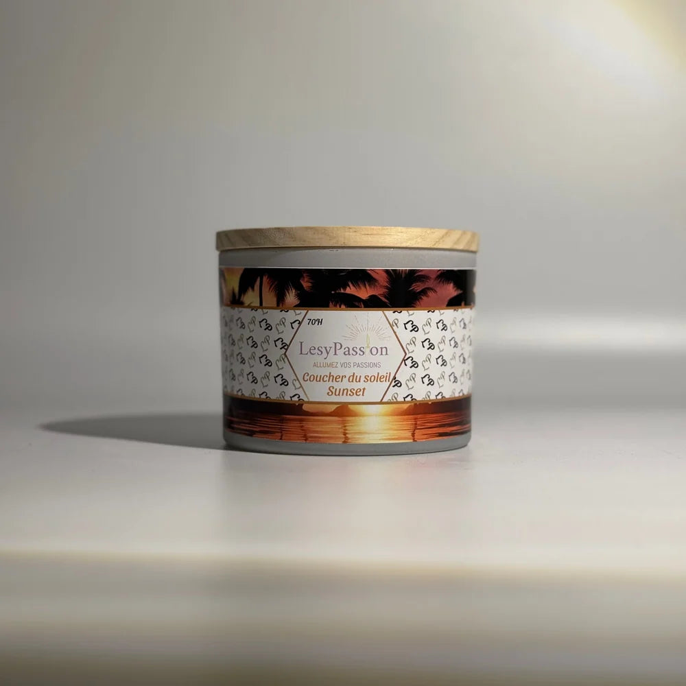 Bougie parfumée grand format 15 oz en pot large gris clair surmonté d'un couvercle en bois clair, dotée d'une étiquette « LesyPassion - Coucher du soleil / Sunset » illustrée d'un coucher de soleil flamboyant sur l'océan avec silhouettes de palmiers, sur fond studio neutre.