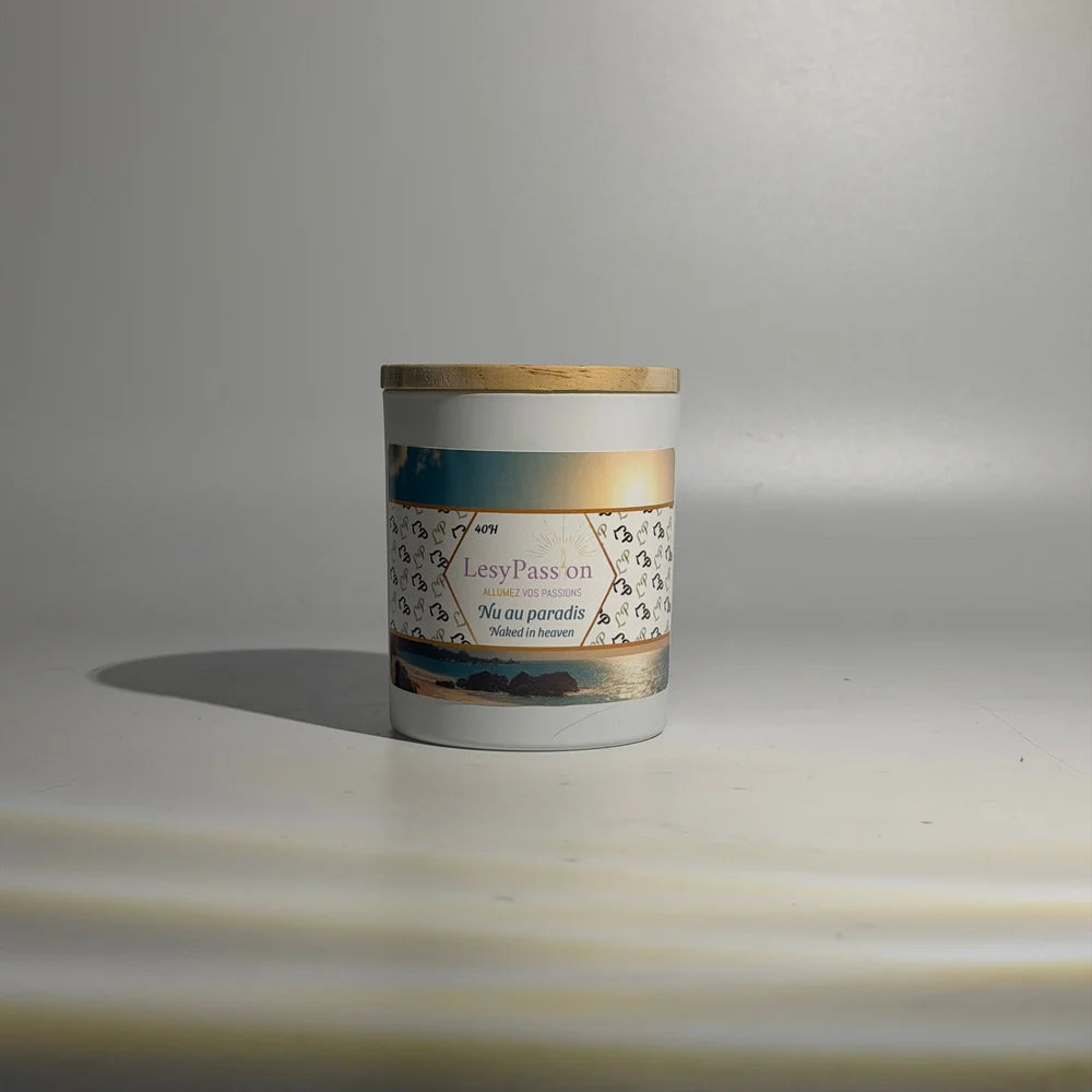 Bougie parfumée format 9 oz (40 h) en pot cylindrique blanc avec couvercle en bois clair, dotée d'une étiquette « LesyPassion - Nu au paradis / Naked in heaven » illustrée d'un coucher de soleil sur une plage tropicale, sur fond studio neutre.