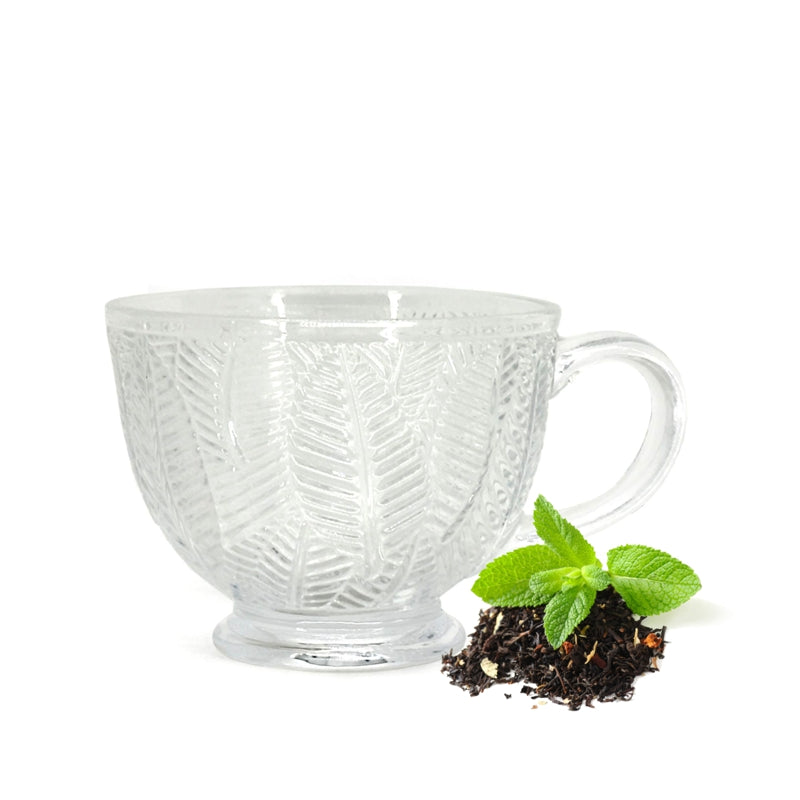 Tasse sur pied en verre moulé transparent ornée de feuillage texturé, avec petit monticule de thé en vrac et feuilles de menthe verte sur fond blanc, Danesco