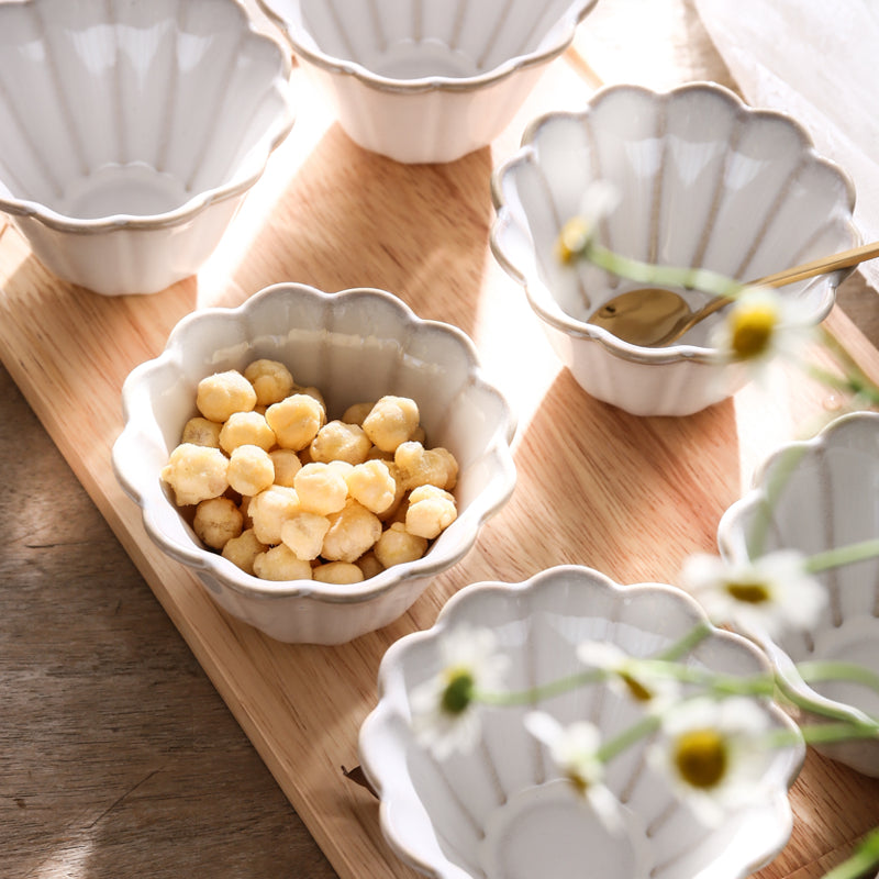 Mise en table lumineuse montrant plusieurs ramequins ondulés en grès blanc sur une planche de bois clair, dont un rempli de petites bouchées et un avec une cuillère à café dorée, décorés de fleurs au premier plan, la petite cuisine de Danesco