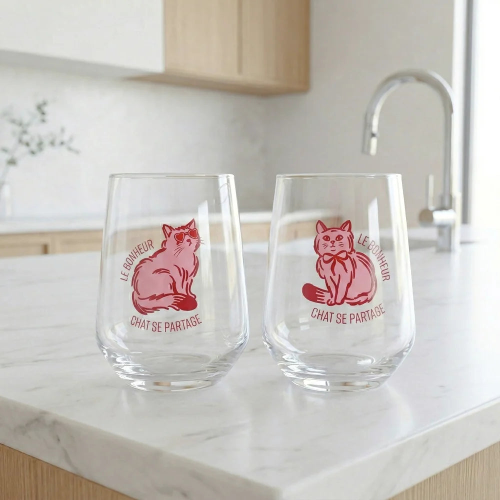 Deux verres sans pied transparents sur un comptoir en marbre, décorés d'illustrations de chats rouges et de l'inscription « Le bonheur chat se partage ».