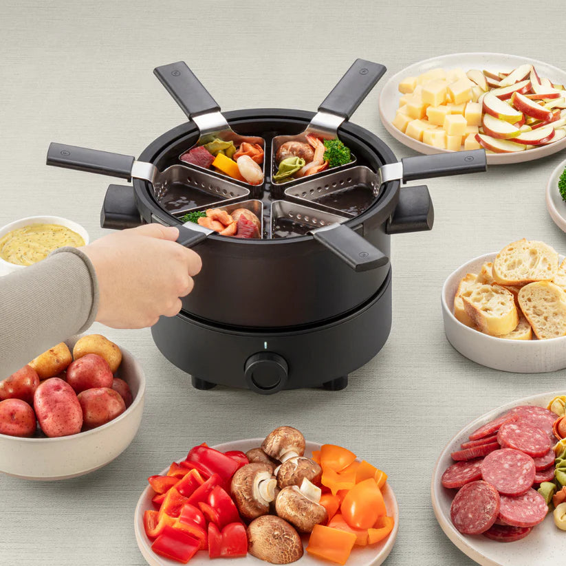 Poêle à fondue électrique noire avec six paniers perforés en cours d'utilisation, entourée de bols de légumes, viandes, charcuteries, pains et fromages.