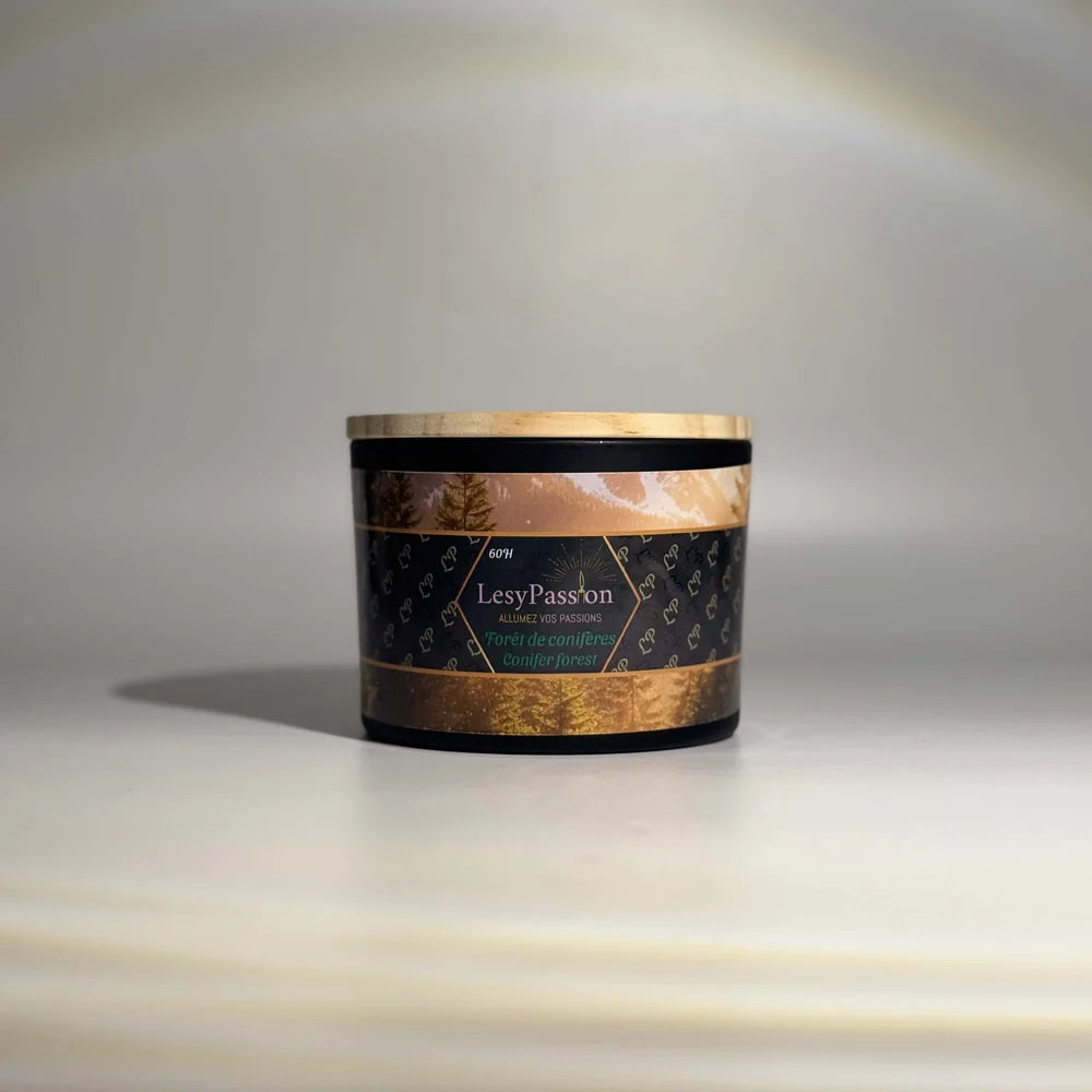 Bougie parfumée grand format (60 h) en pot large noir mat surmonté d'un couvercle en bois clair, dotée d'une étiquette « LesyPassion - Forêt de conifères / Conifer forest » ornée d'illustrations de sapins dorés, sur fond studio neutre.