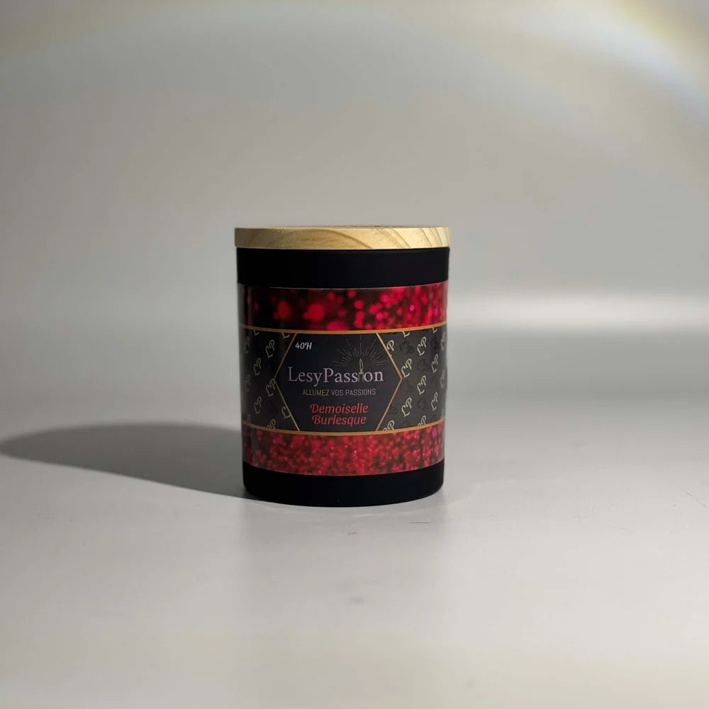 Bougie parfumée format 9 oz (40 h) en pot cylindrique noir mat avec couvercle en bois clair, ornée d'une étiquette « LesyPassion - Demoiselle Burlesque » aux motifs de paillettes rouge rubis, sur fond studio neutre.