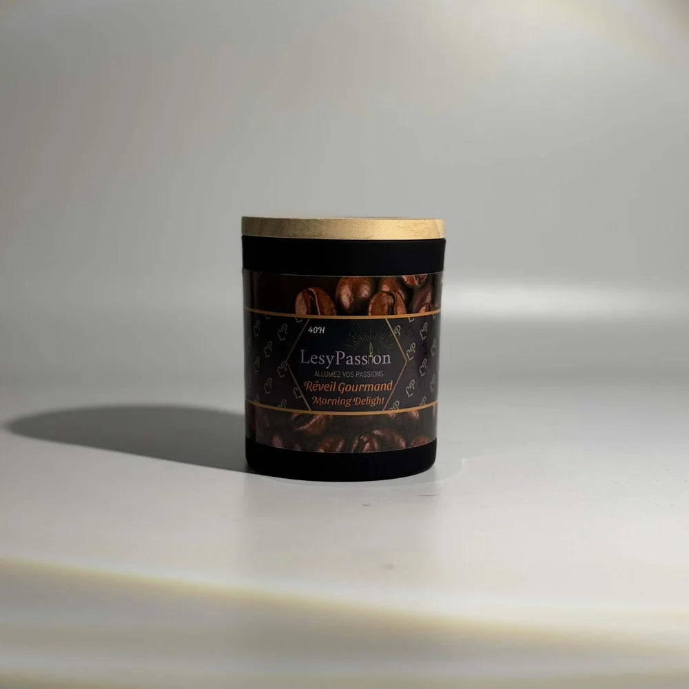 Bougie parfumée format 9 oz (40 h) en pot cylindrique noir mat avec couvercle en bois clair, dotée d'une étiquette « LesyPassion - Réveil Gourmand / Morning Delight » illustrée de grains de café torréfiés, sur fond studio neutre.