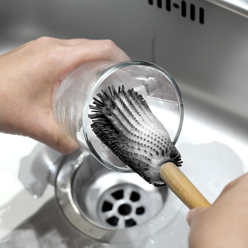 Démonstration de nettoyage : une main tient un verre à eau tandis qu'un goupillon à vaisselle en silicone gris avec manche en bois frotte l'intérieur avec de la mousse de savon, au-dessus de la bonde d'un évier en acier inoxydable