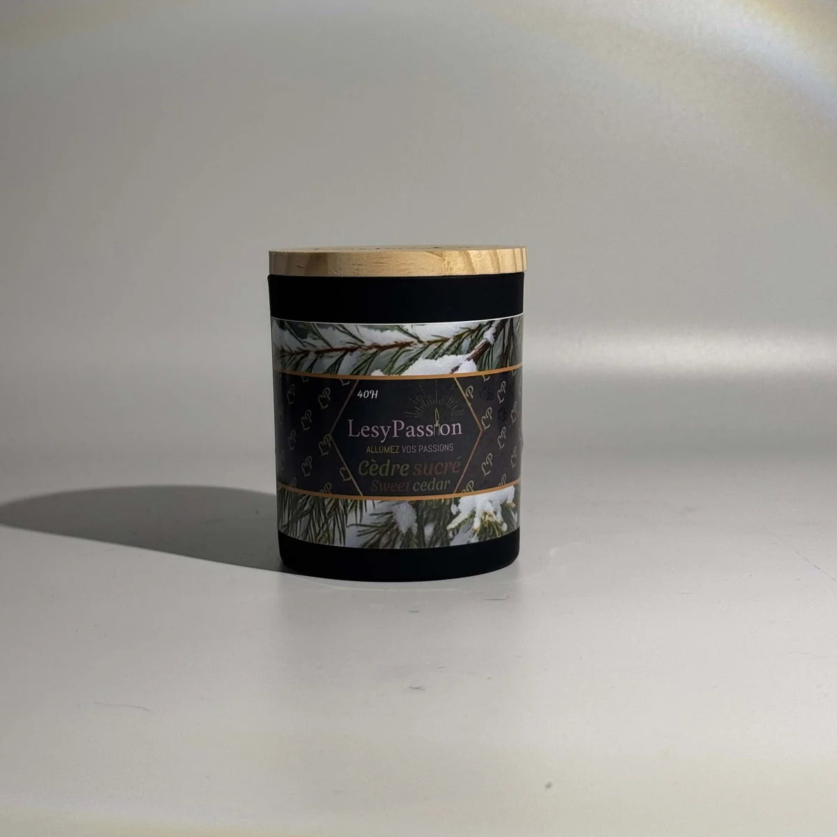 Bougie parfumée format moyen (40 h) en pot cylindrique noir mat avec couvercle en bois clair, dotée d'une étiquette « LesyPassion - Cèdre sucré / Sweet cedar » illustrée de branches de conifères enneigées, sur fond studio épuré.