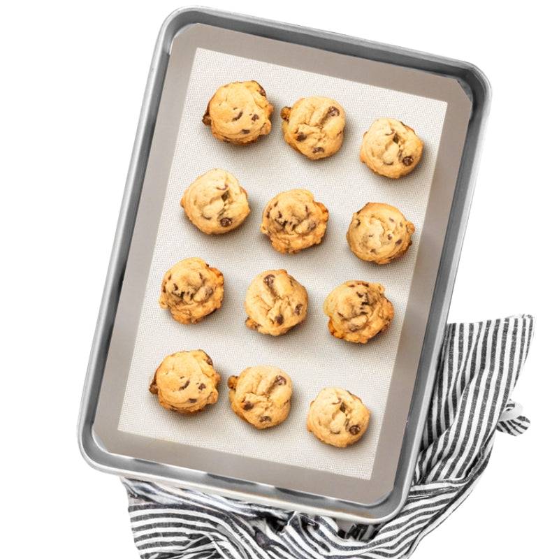 Mise en situation de biscuits aux pépites de chocolat dorés sur un tapis de cuisson réutilisable en silicone à bordure grise, avec linge rayé sur fond blanc, de la compagnie Danesco 